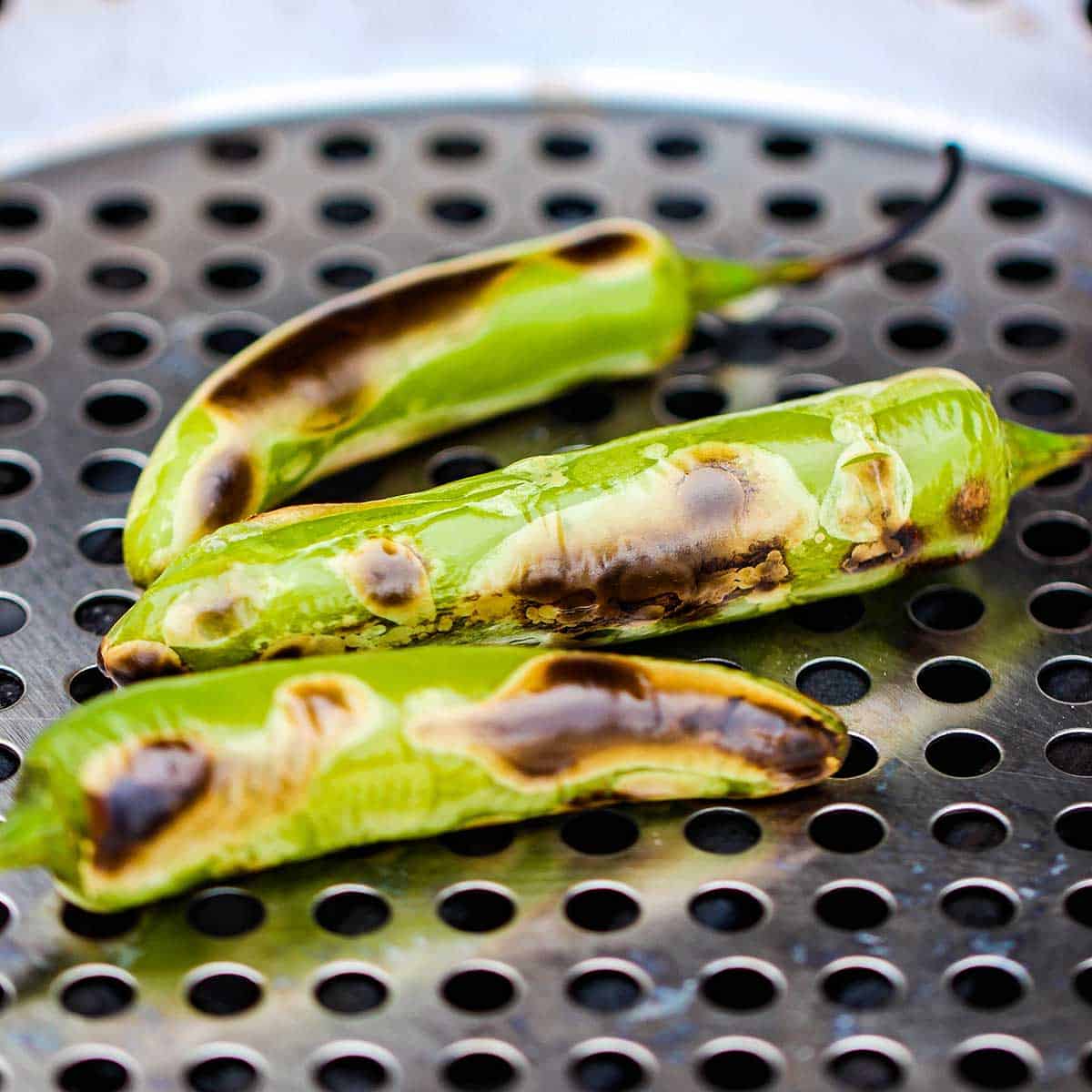 Three whole serrano peppers that are blistered and are resting on a metal grill pan on a gas grill.