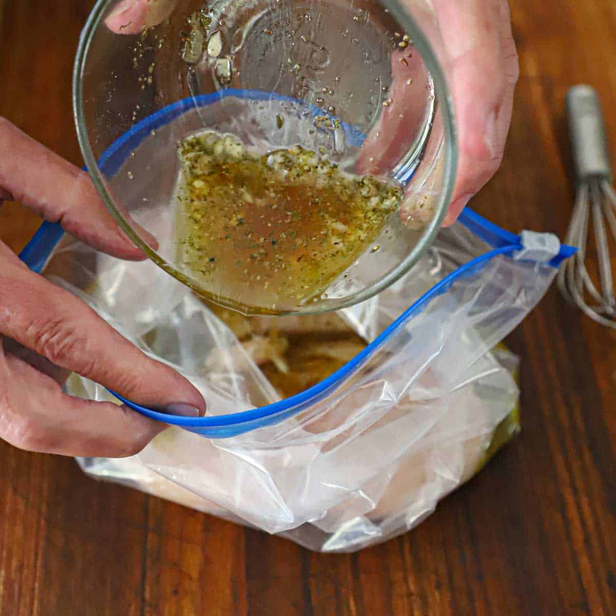 A person pouring a marinade from a small glass bowl into a plastic baggies that contains two uncooked chicken breasts.