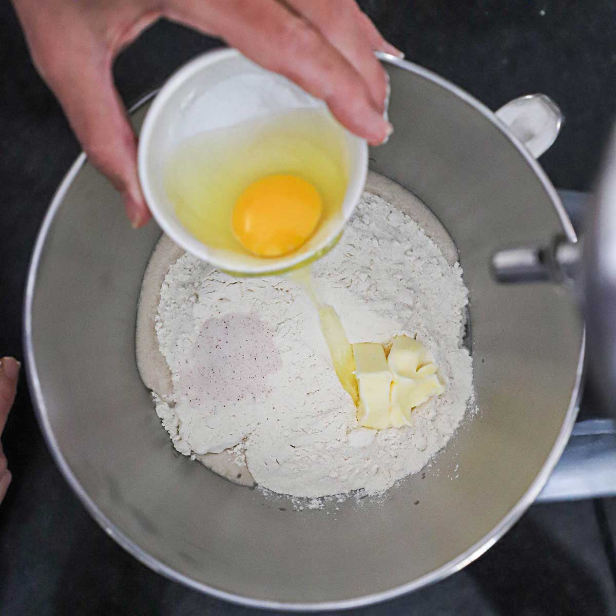 A person transferring a whole egg from a small white bowl into the bowl of a stand mixer that is filled with foamy yeast water, flour, salt, and softened butter.