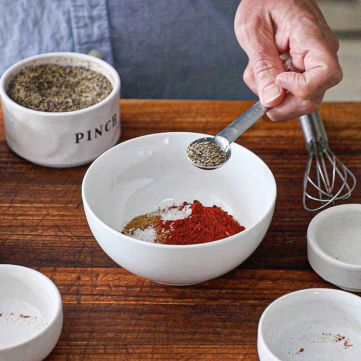 A person holding a half teaspoon filled with ground black pepper over a white bowl filled with chili powder, cumin, and other seasonings for homemade taco seasoning.