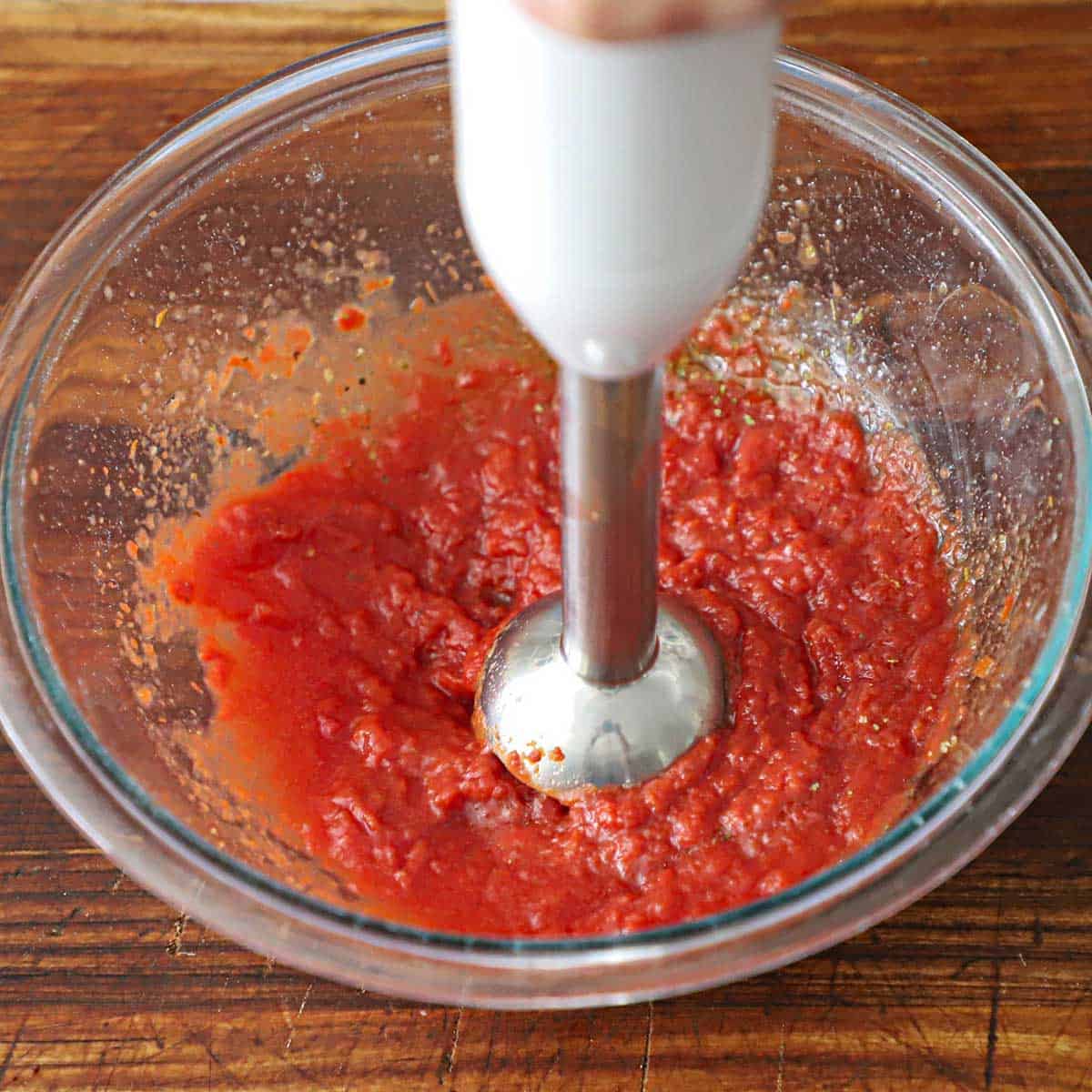 An immersion blender blending ground tomatoes in a glass bowl sitting on a wooden cutting board.