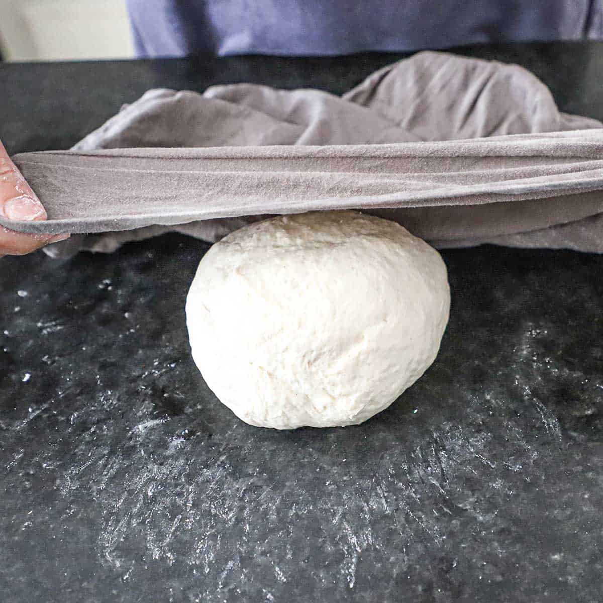 A person placing a damp grey napkin over a round ball of New York-style pizza dough on a black marble countertop.