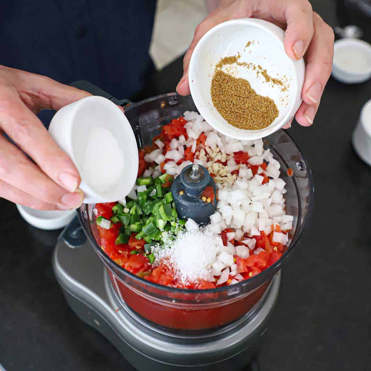 A person adding sugar and dried cumin into a food processor that is filled with all of the ingredients for making restaurant-style salsa including chopped onion, jalapeño, garlic, tomatoes, and seasonings.