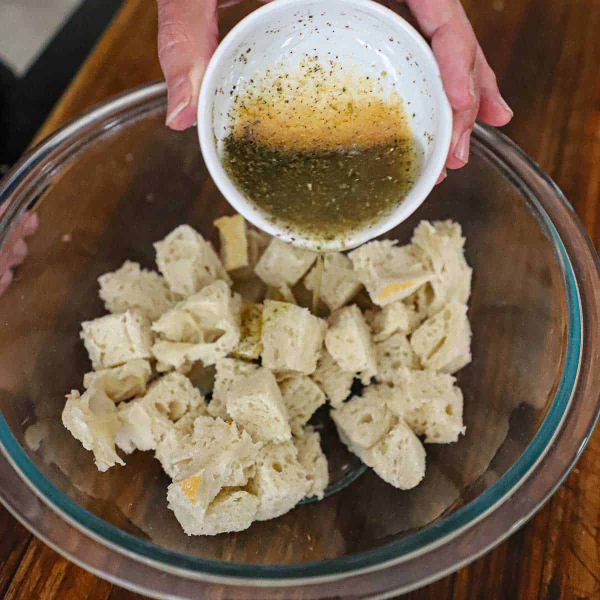 A person pouring seasoned oil over fresh cubes of bread that are sitting in a glass bowl on a cutting board.
