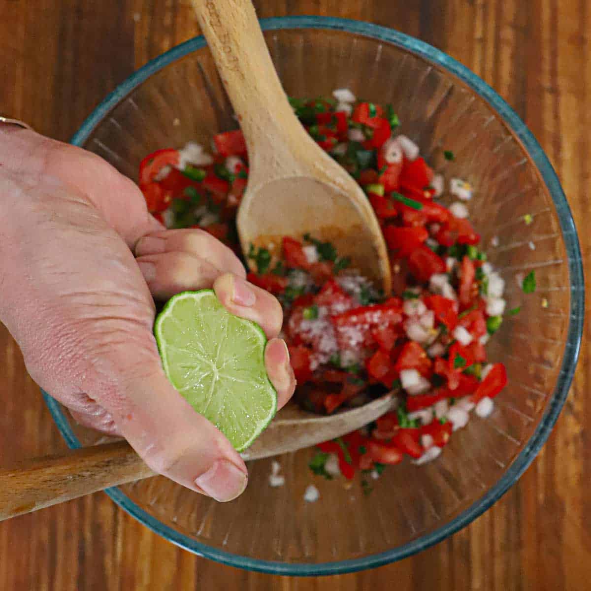 A person squeezing a fresh lime over a glass bowl filled with freshly made pico de gallo.