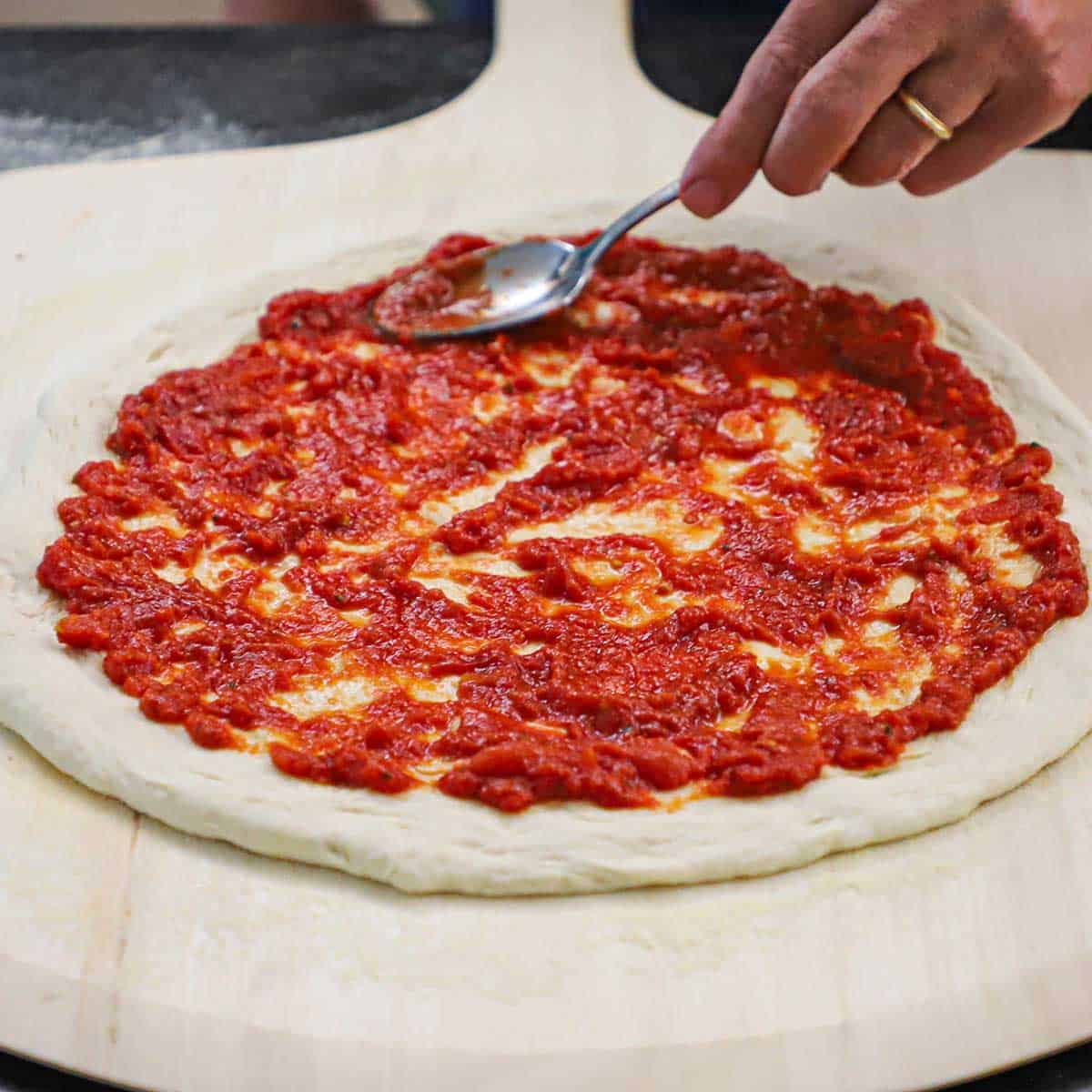 A person using a spoon to spread pizza sauce on a circular pizza dough that is sitting on a large pizza wheel.