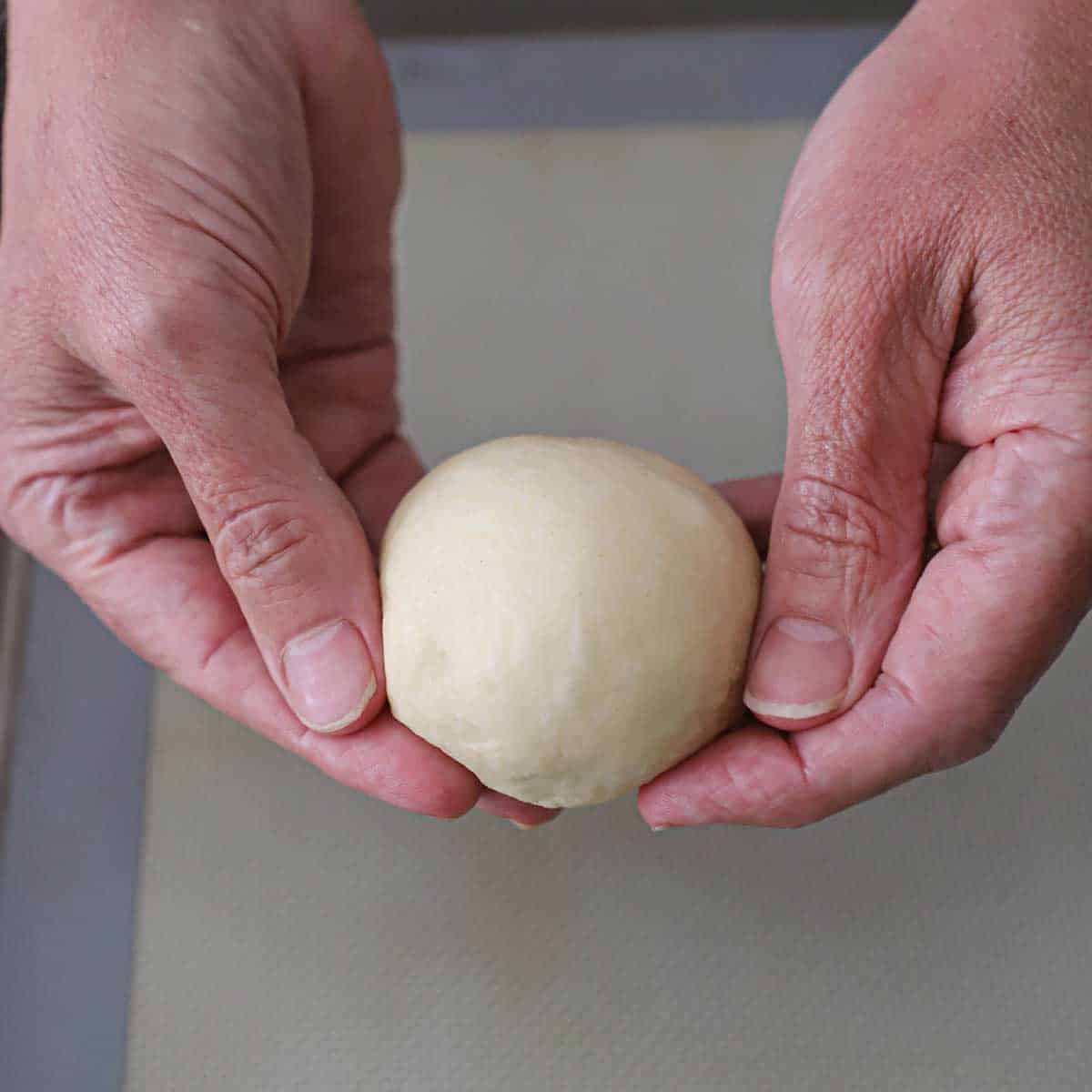 A person holding a smooth ball of hamburger bun dough in his hands.