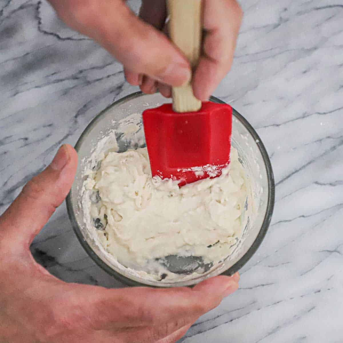 A person using a red rubber spatula to incorporate high-gluten flour into a small bowl filled a water and yeast mixture to create poolish.