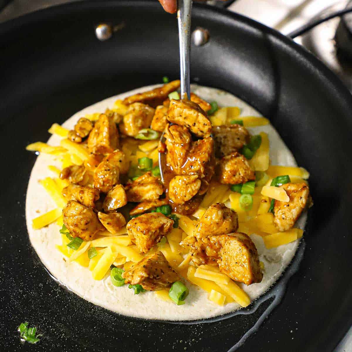 A person placing a spoonful of cooked taco chicken pieces in the center of more taco chicken that is on a tortilla with cheese in a skillet simmering with oil.
