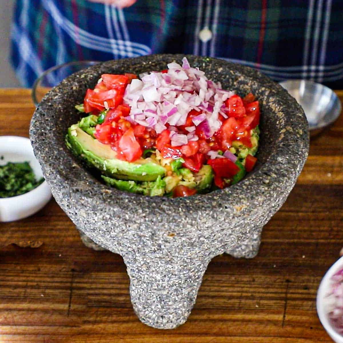 A molcajete sitting on a wooden cutting board that is filled with chunks of avocado, chopped tomatoes, and chopped red onion.