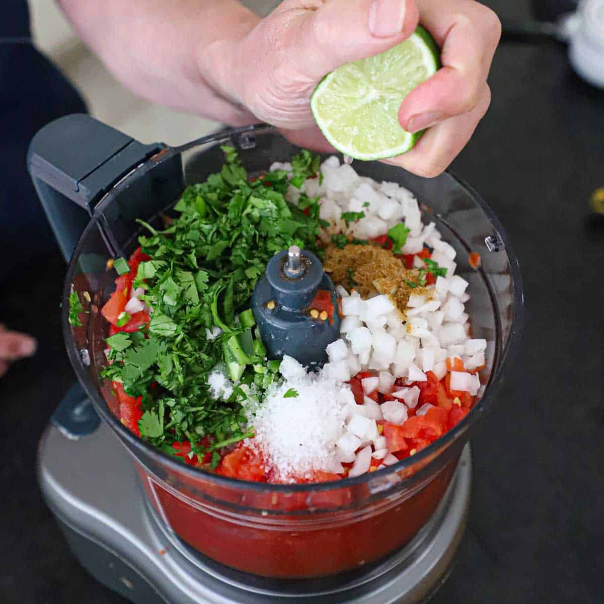 A person squeezing a half of a lime into the bucket of a food processor that is filled with the ingredients for restaurant-style salsa.
