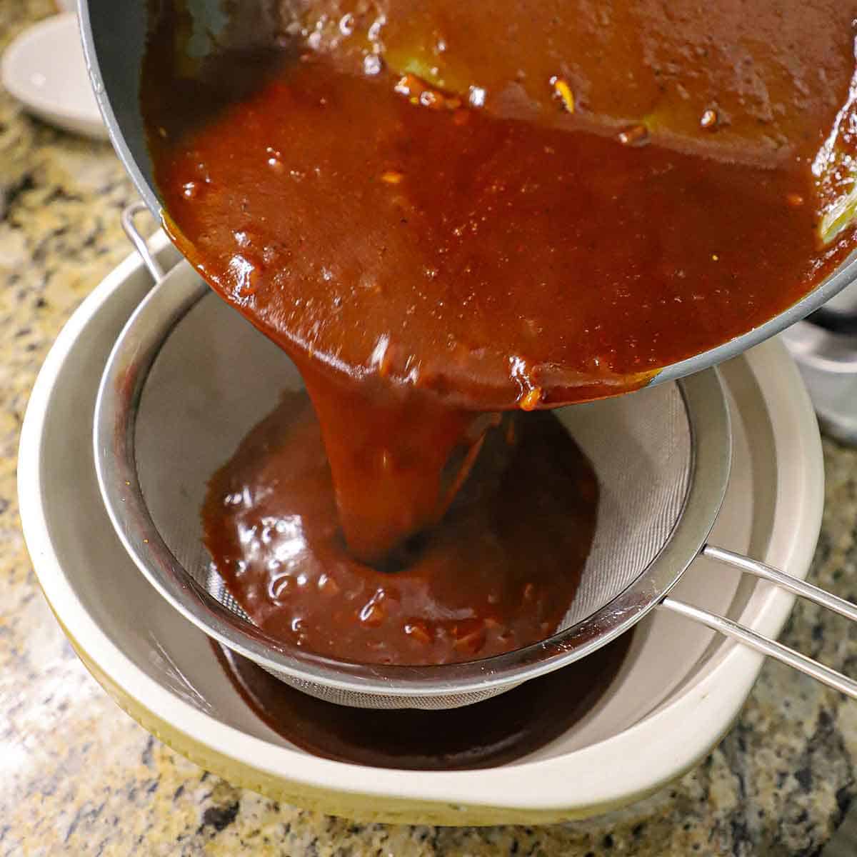 A person pouring homemade BBQ sauce through a sieve that is resting over a ceramic bowl on a marble counter.