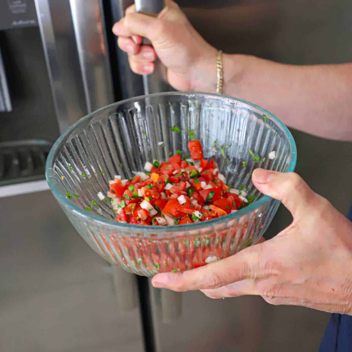 A person holding a glass bowl filled with freshly made pico de gallo and the other hand on the handle of a stainless steel refrigerator.