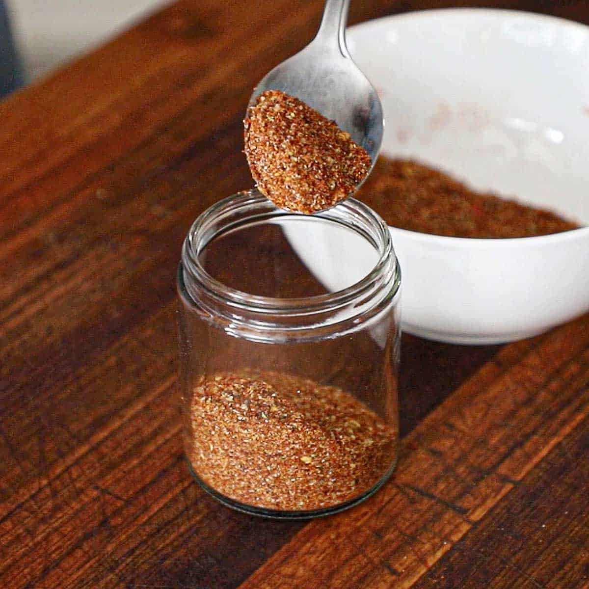 A person holding a spoonful of homemade taco seasoning over a small glass jar that is partially filled with the taco seasoning.