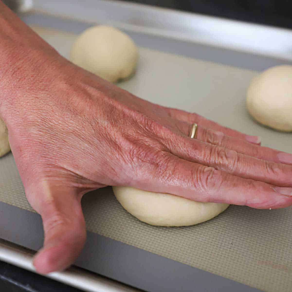 A person using the palm of his hand to gently flatten a ball of hamburger bun dough on a sheet pan lined with a silicone matt.