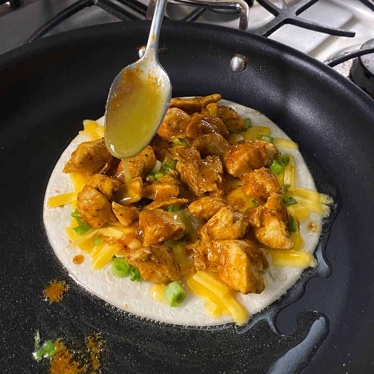 A person spooning green enchilada sauce over the ingredients on the inside of a chicken quesadilla simmering in a hot skillet with oil in it.