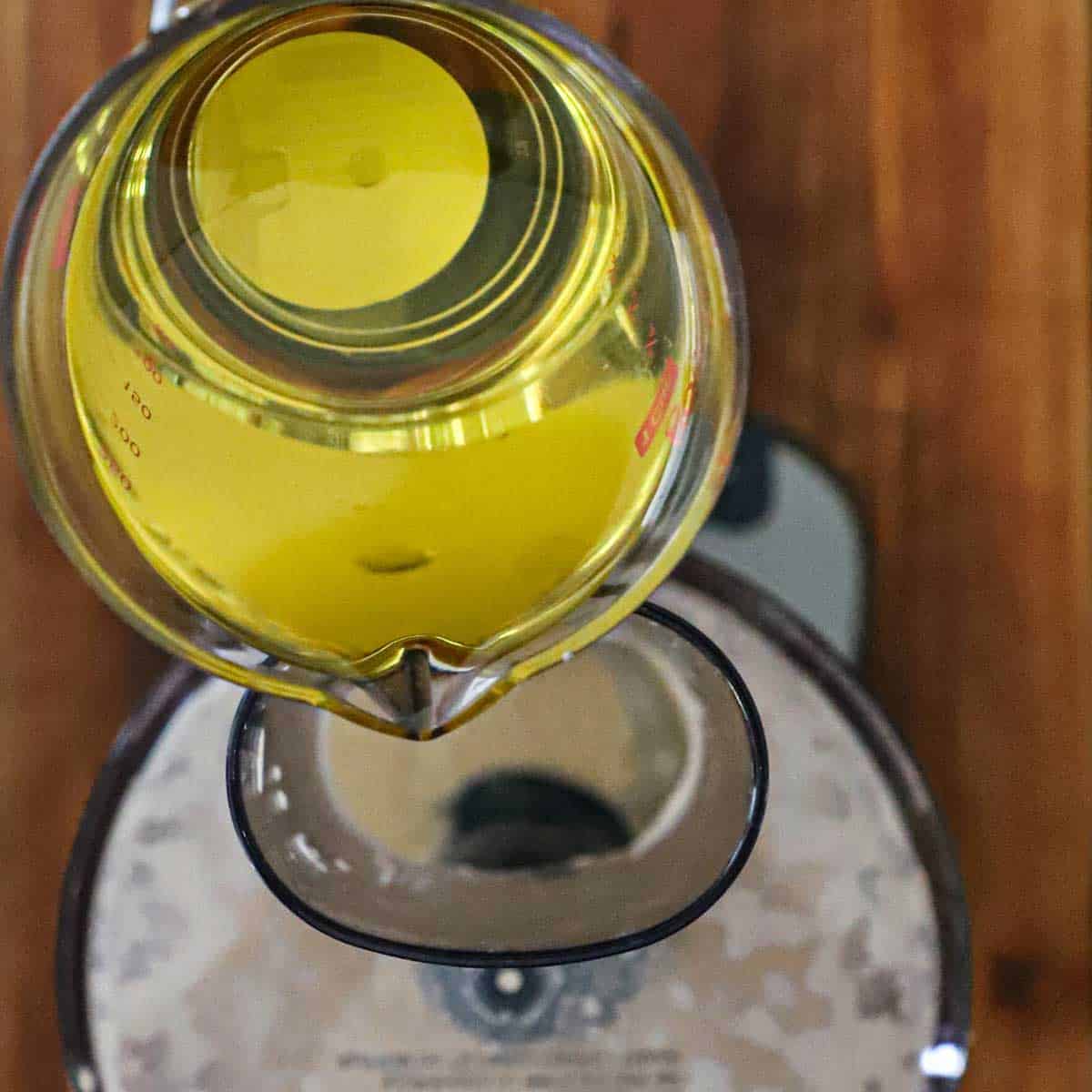 A person pouring olive oil from a glass measuring cup through the tube of a food processor in the bucket that is filled with the ingredients for Caesar dressing.