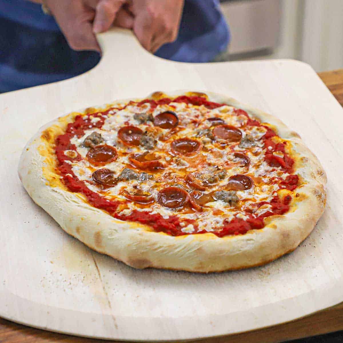 A person holding a freshly baked homemade New York-style pizza on a large pizza wheel.