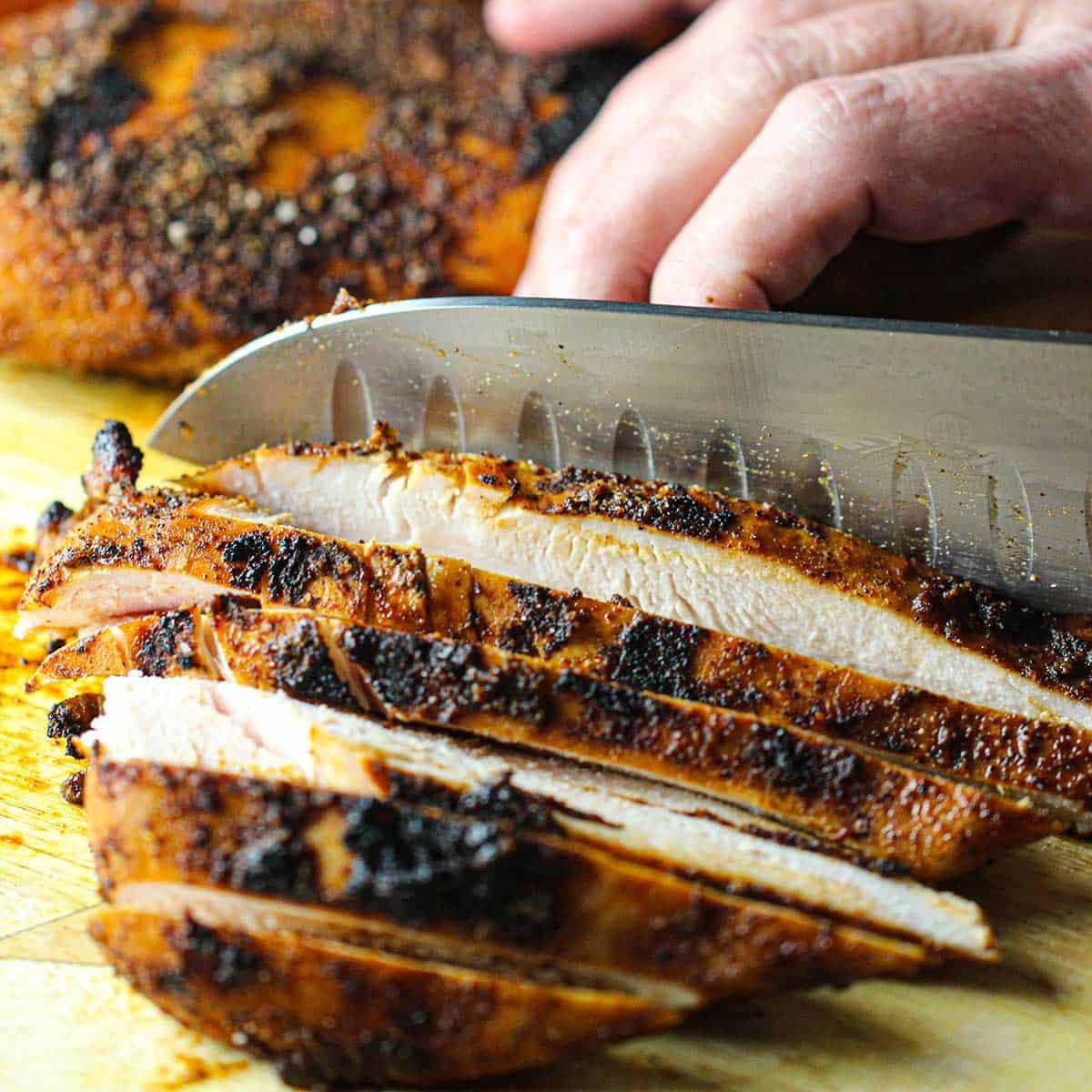 A person using a large chef's knife to slice grilled chicken fajita meat into strips on a wooden cutting board.