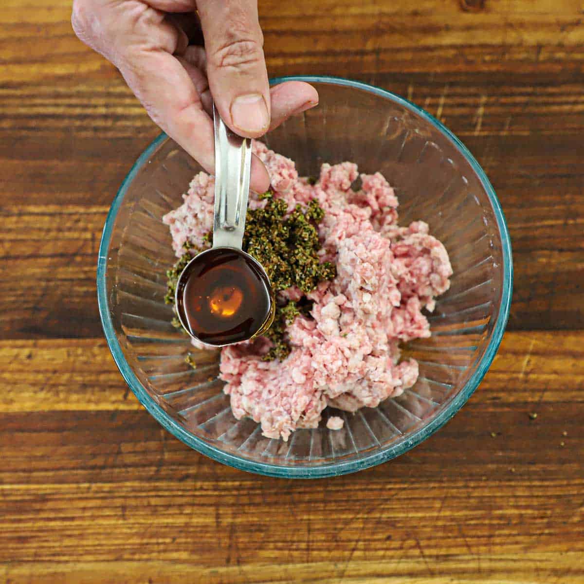 A person holding a tablespoon of honey over a glass bowl filled with uncooked ground pork topped with dried seasonings.