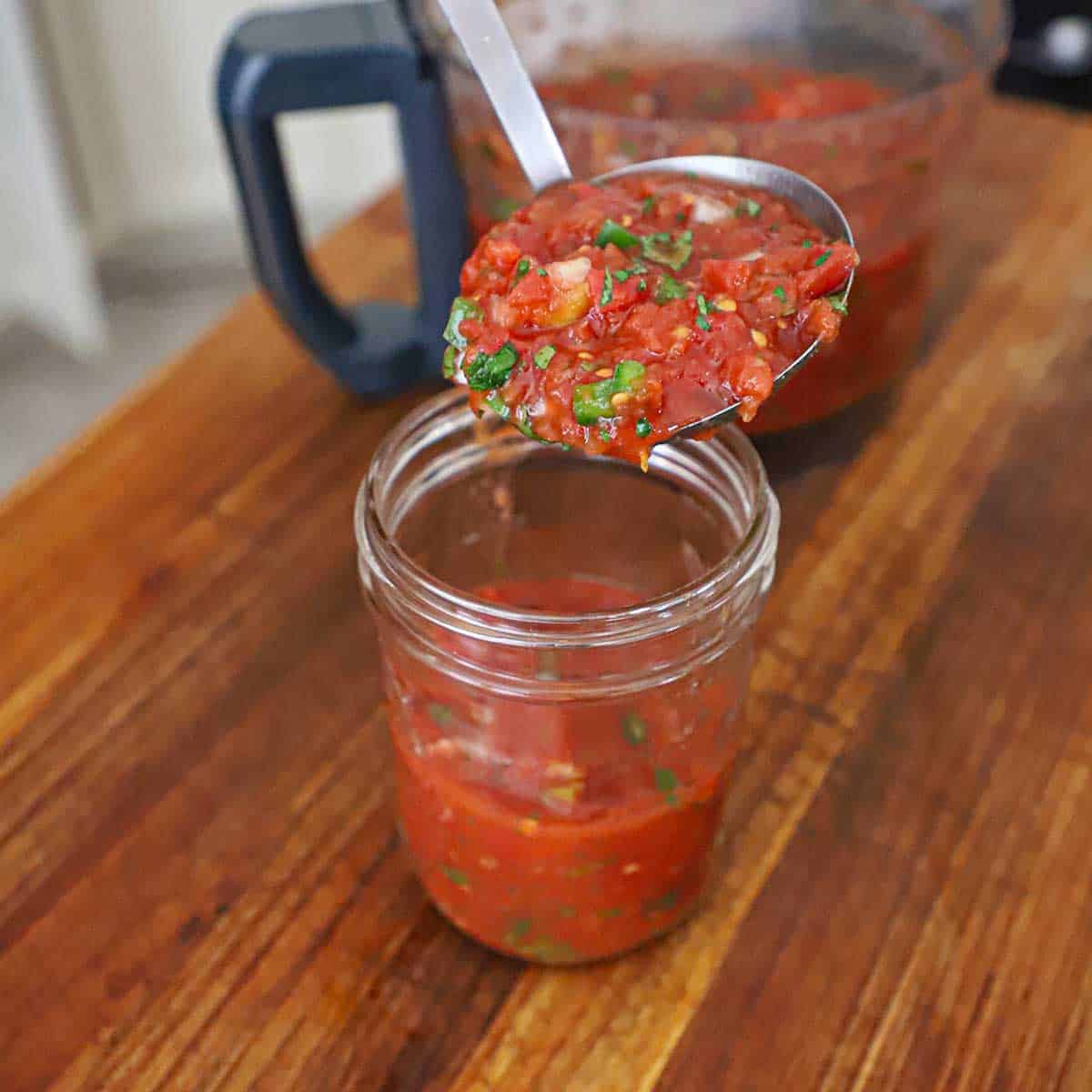 A person using a small metal ladle to transfer restaurant-style salsa into a small glass jar that is resting on a cutting board with a food processor holding the salsa in the background.