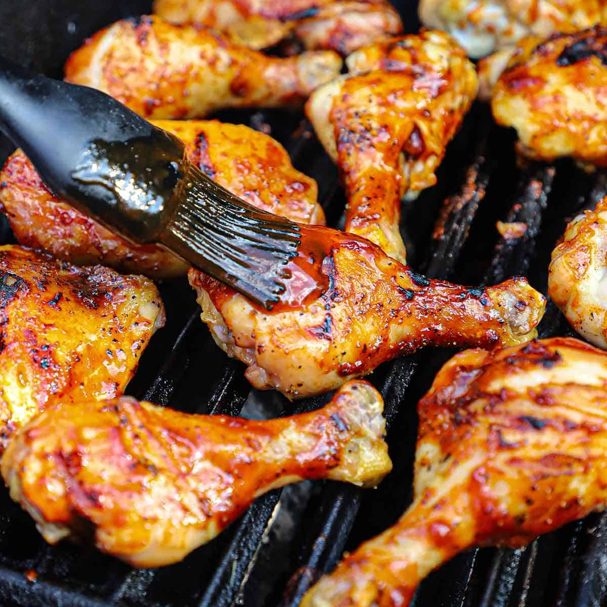 A person brushing homemade BBQ sauce onto a grilled chicken leg that is being cooked on a gas grill with other grilled BBQ chicken pieces.