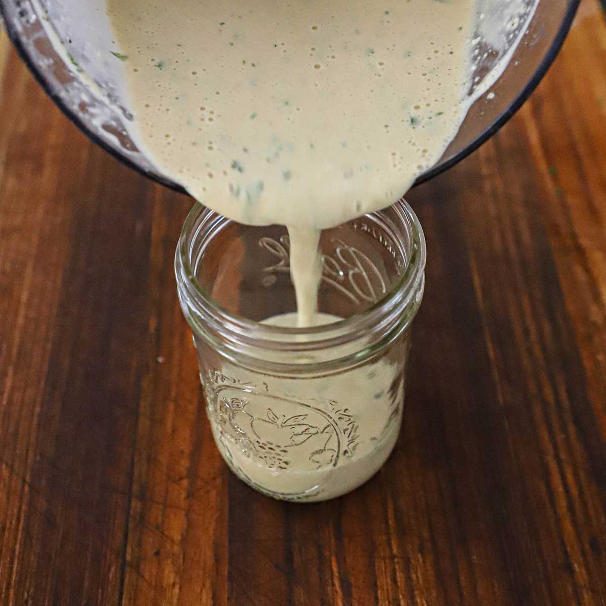 A person pouring homemade Caesar dressing from a glass bowl into a glass jar that is sitting on a wooden cutting board.