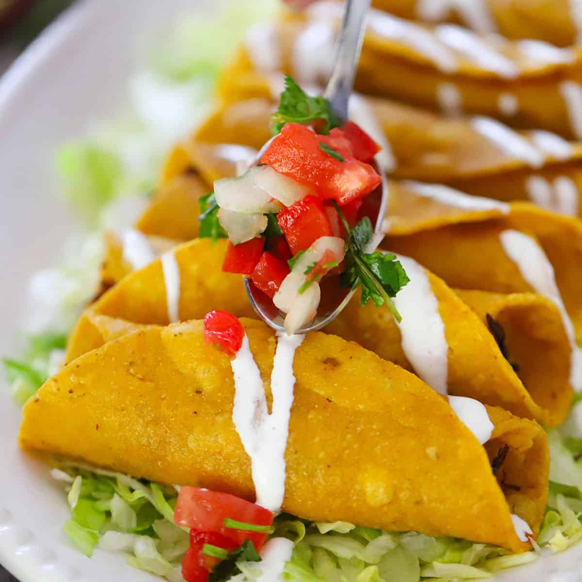 A person using a small spoon to top a row of crispy chicken flautas of homemade pico de gallo.