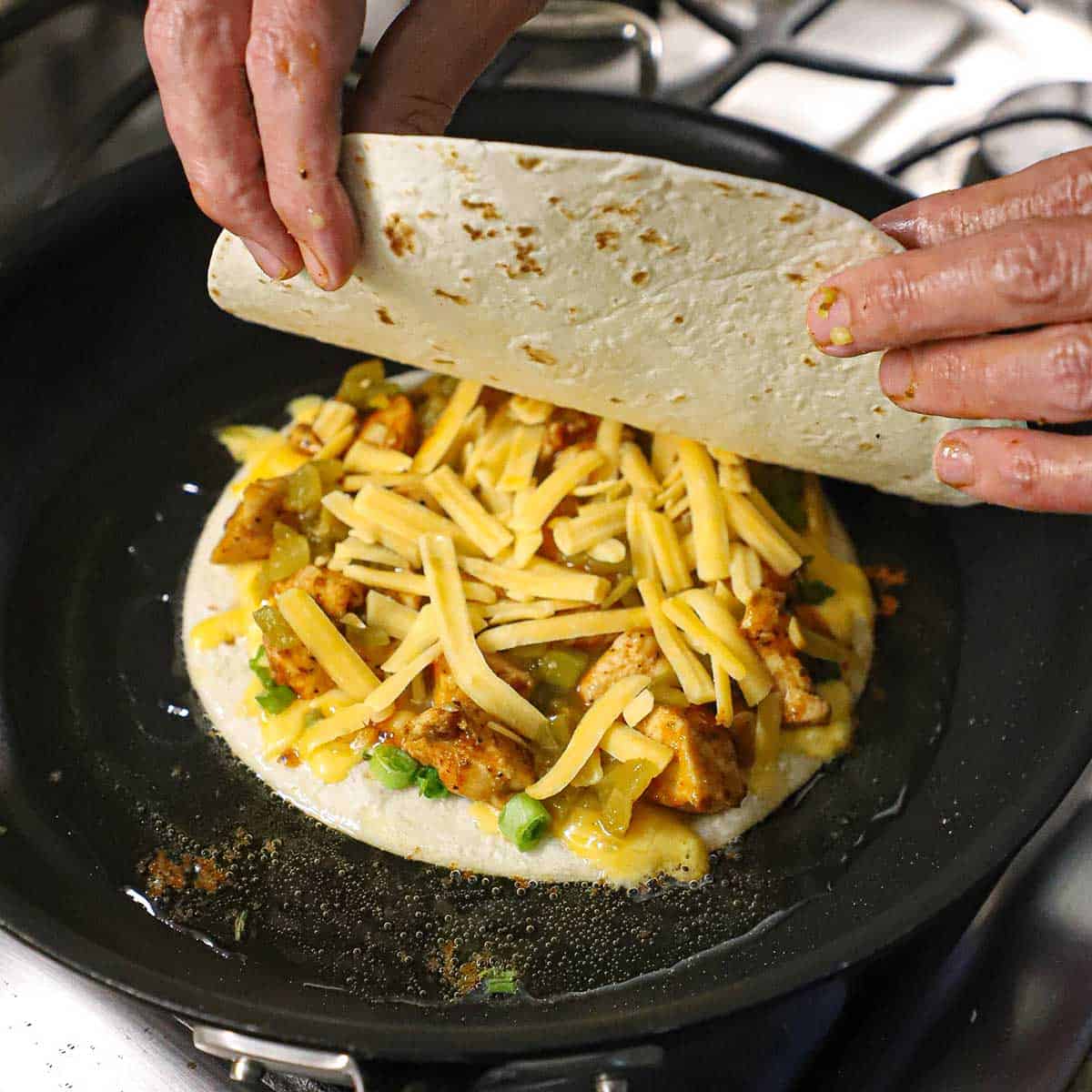 A person placing a flour tortilla over the top of a chicken quesadilla that is being simmering in hot oil in a large skillet.