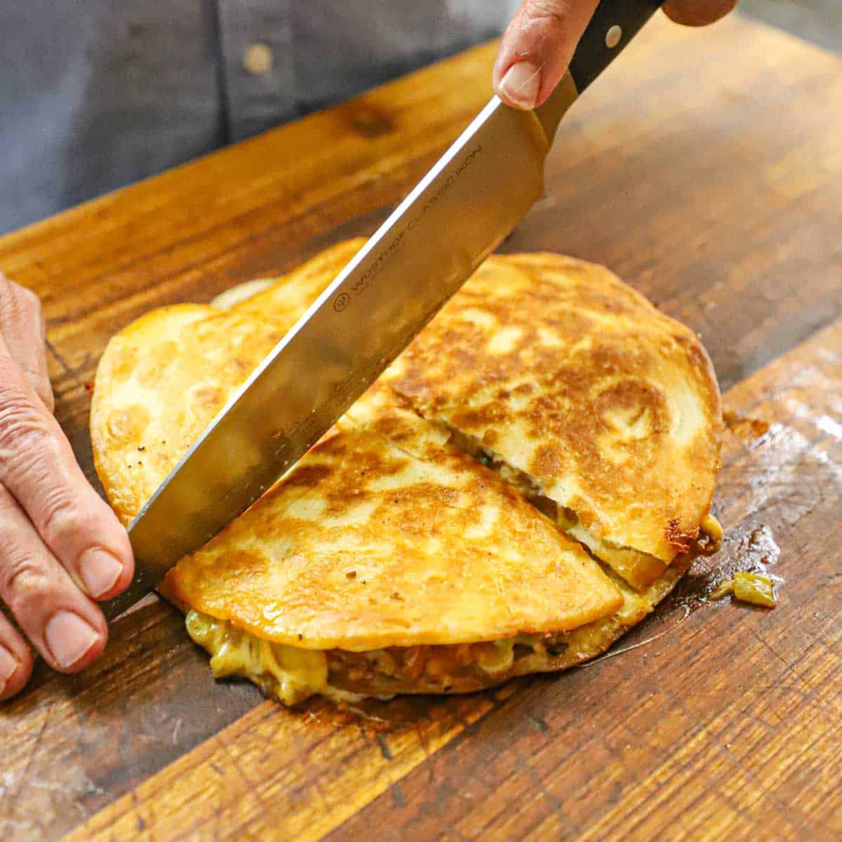 A person using a large chef's knife to cut a crispy chicken quesadilla into quarters on a wooden cutting board.