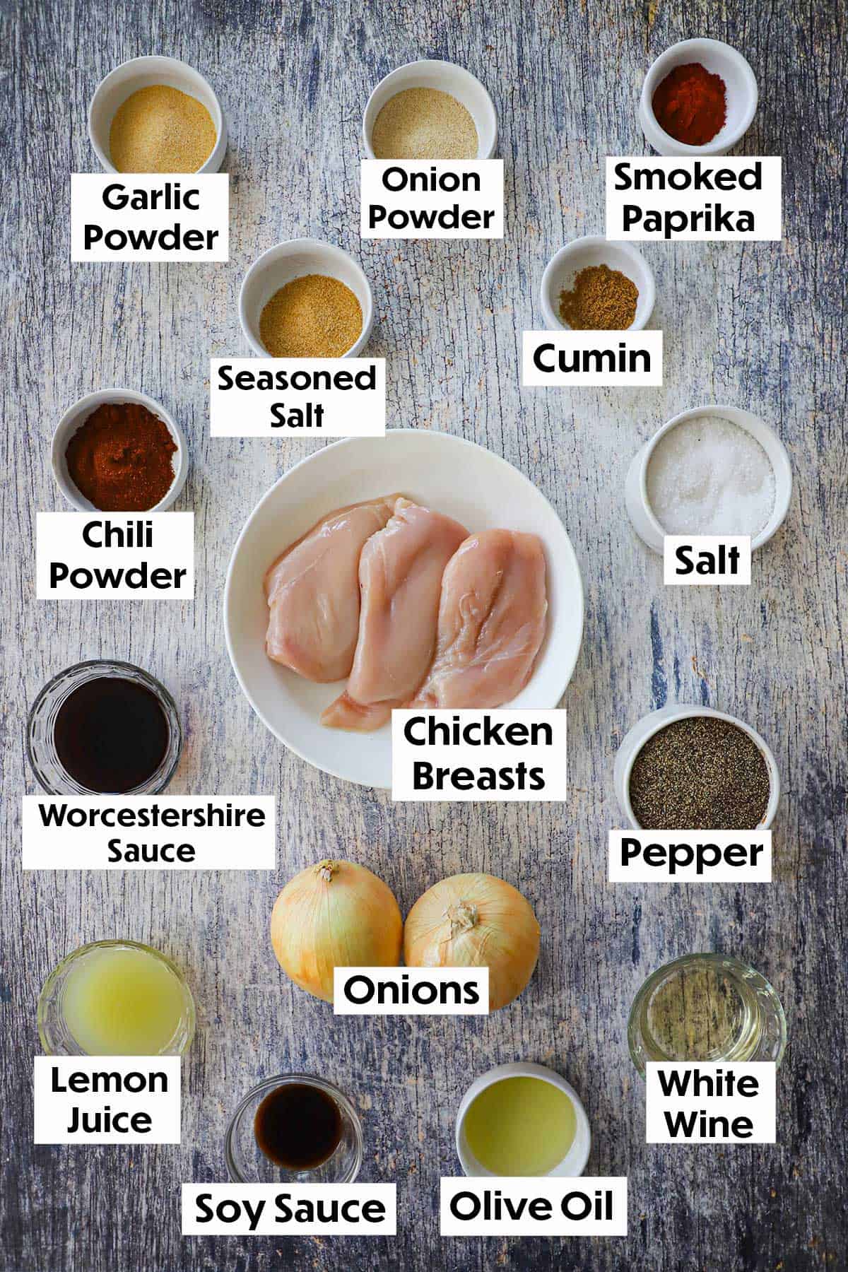 An arrangement of ingredients for Tex-Mex chicken fajitas on a grey wooden background including seasonings, uncooked chicken breasts, Worcestershire sauce, onions, lemon juice, soy sauce, and white wine.