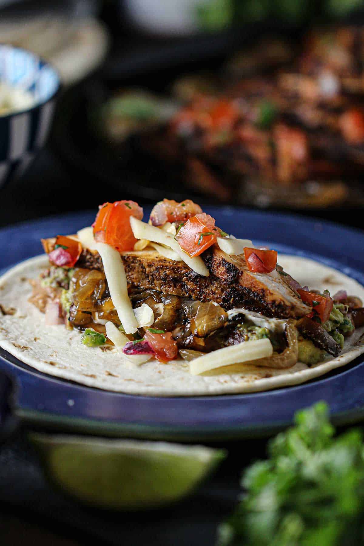 An open Tex-Mex chicken fajita topped with grated Monterrey Jack cheese and pico de gallo resting on a blue dinner plate surround by lime wedges and sprigs of cilantro.