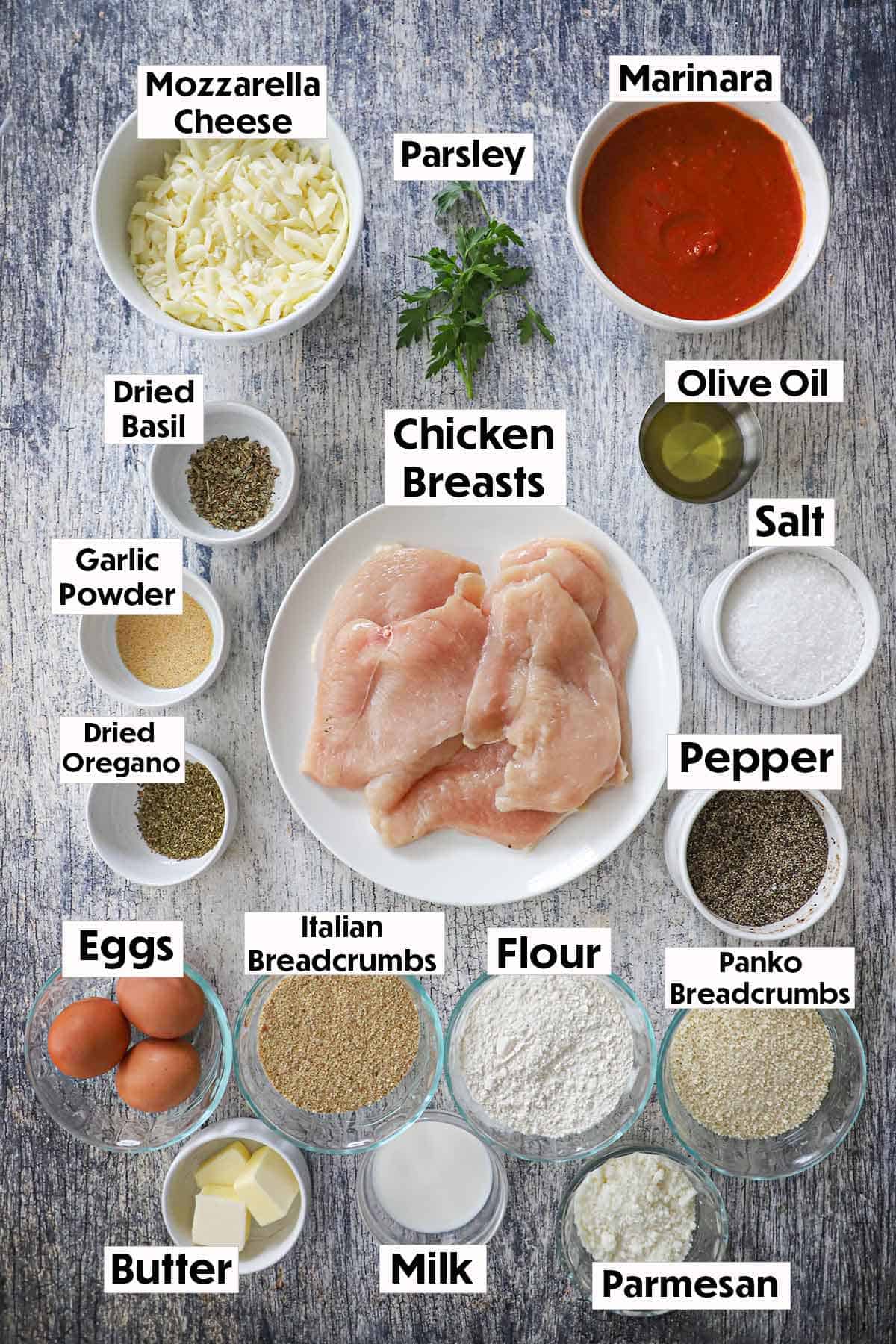 An arrangement of ingredients for chicken Parmesan (Parmigiana) on a grey wooden background including uncooked chicken breasts, marinara sauce, cheese, spices, herbs, breadcrumbs, eggs, butter, milk, and oil.