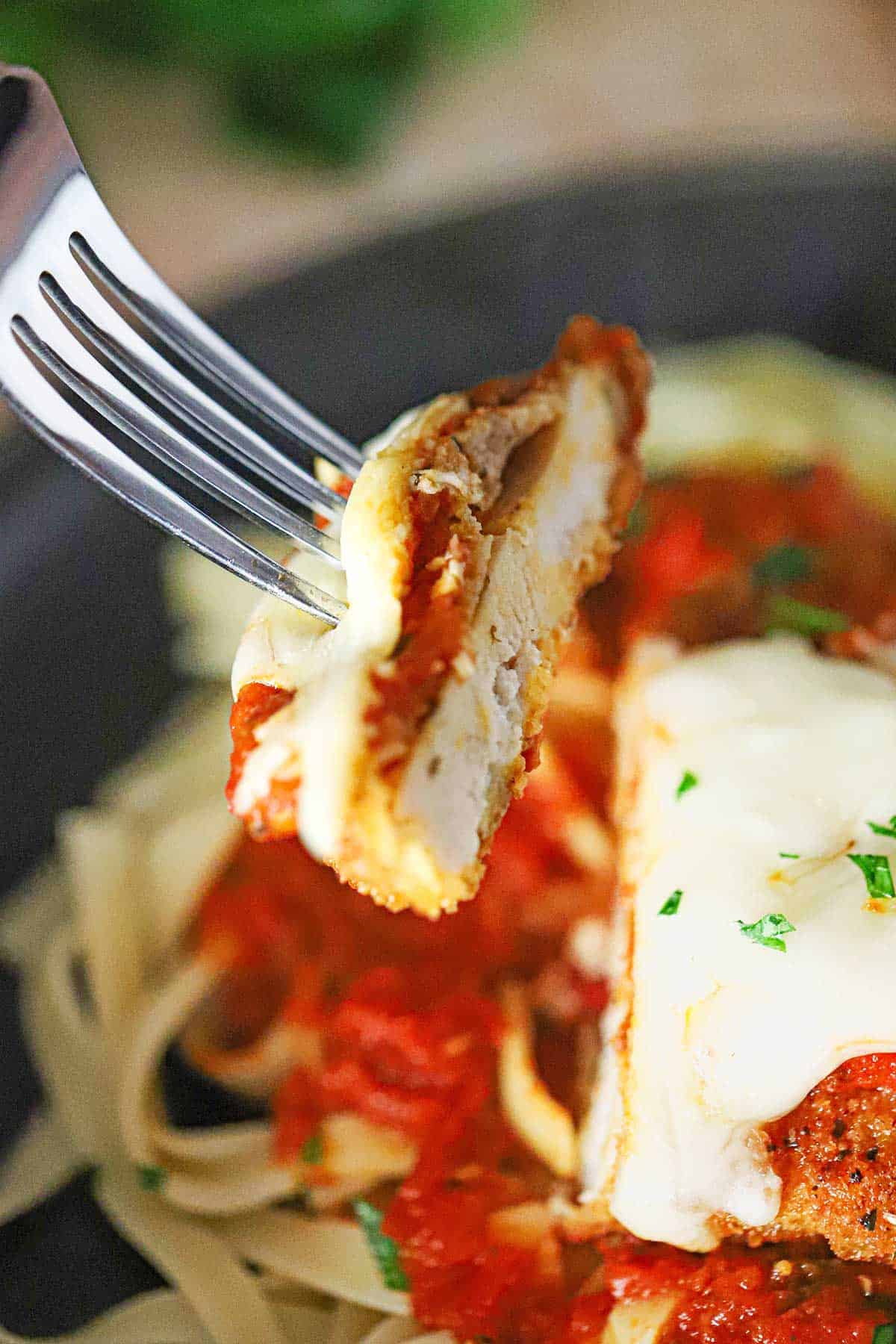 A person using a fork to hold up a cut out piece of chicken Parmesan (Parmigiana) over a plate of the same including marinara sauce and linguine.