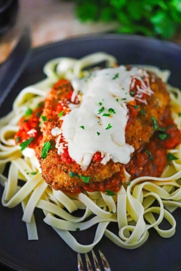 A black dinner plate filled with a serving of chicken Parmesan (Parmigiana) resting on a bed of cooked linguine and marinara sauce.