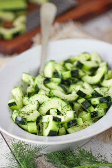 A white oval serving dish filled with sliced cucumber and dill salad with sprigs of fresh dill in front of the bowl.