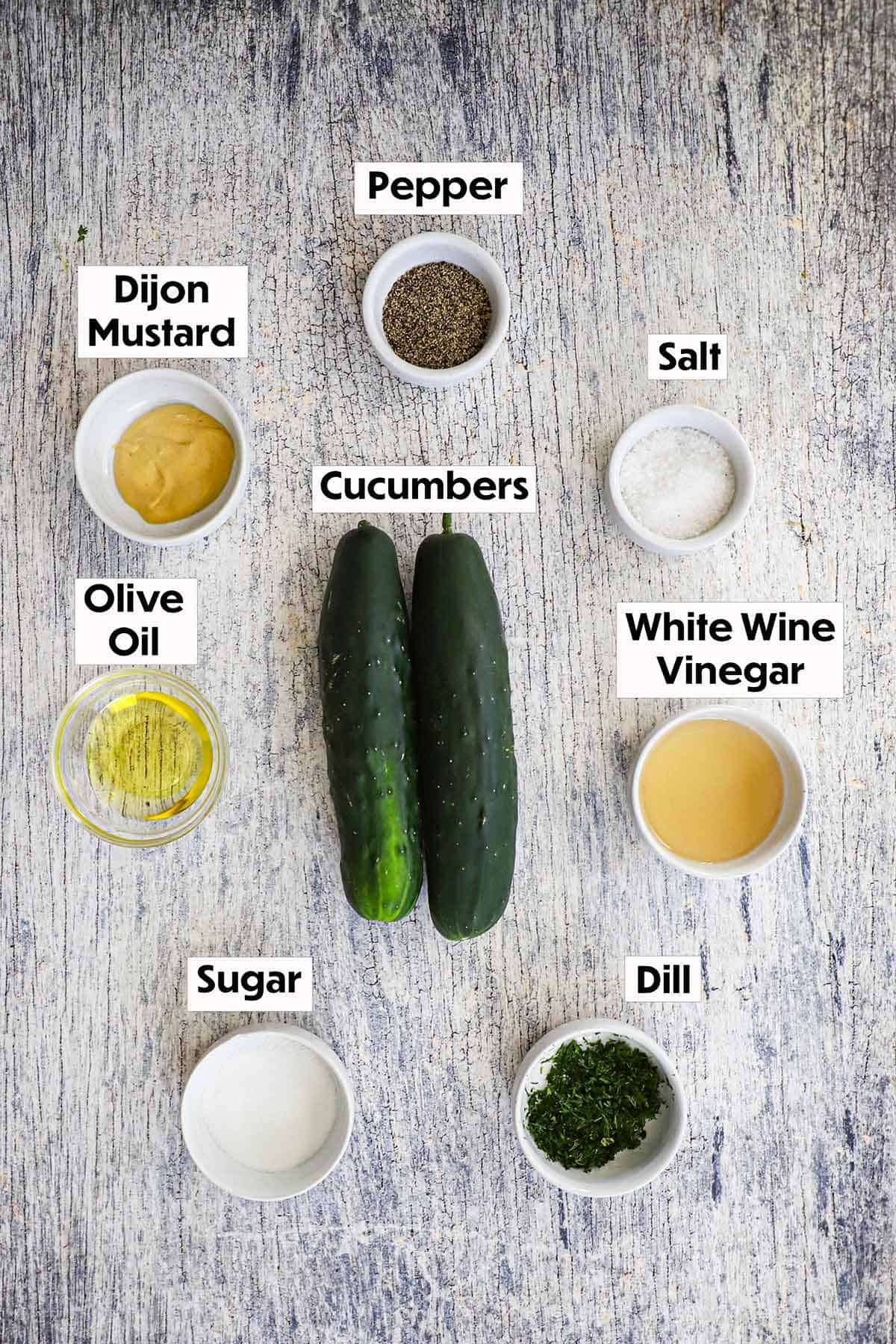 An arrangement of ingredients for cucumber and dill salad on a wooden grey background including two cucumbers, Dijon, oil, vinegar, sugar, chopped dill, salt, and pepper.