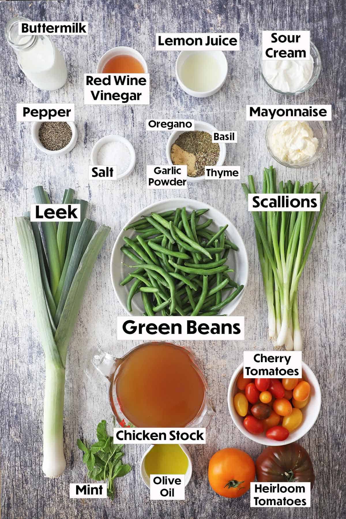 An arrangement of ingredients for green bean and tomato salad on a wooden grey background including fresh green beans, a leek, scallions, buttermilk, sour cream, mayonnaise, vinegar, spices, chicken stock, tomatoes, and herbs.