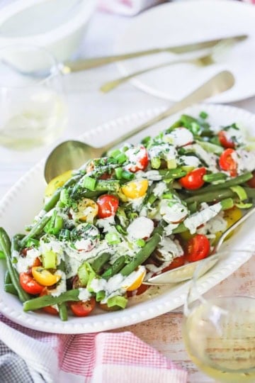 An oval white platter filled with a large serving of green bean and tomato salad topped with a scallion buttermilk dressing, scallions, and chopped fresh herbs.