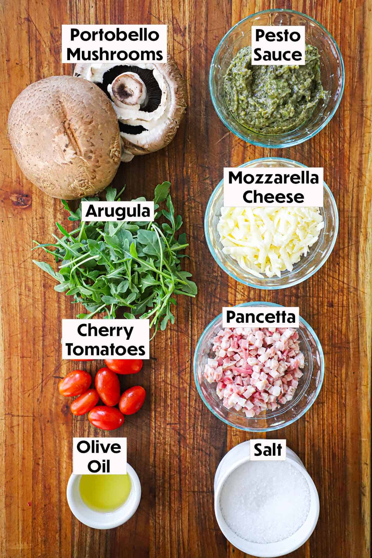 An arrangement of ingredients for portobello BLT pizzas on a wooden background including portobello mushroom caps, pesto, mozzarella cheese, arugula, pancetta, cherry tomatoes, oil, and salt.