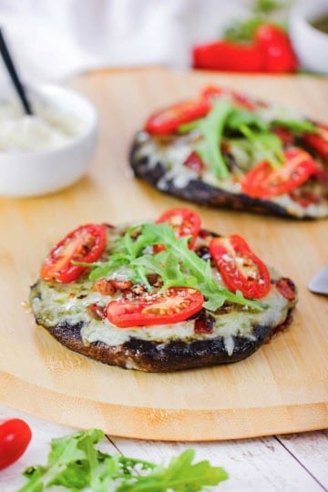 Two portobello BLO pizzas resting on a wooden pizza peel with both pizzas topped with melty Fontina cheese, sliced cherry tomatoes, and arugula leaves.