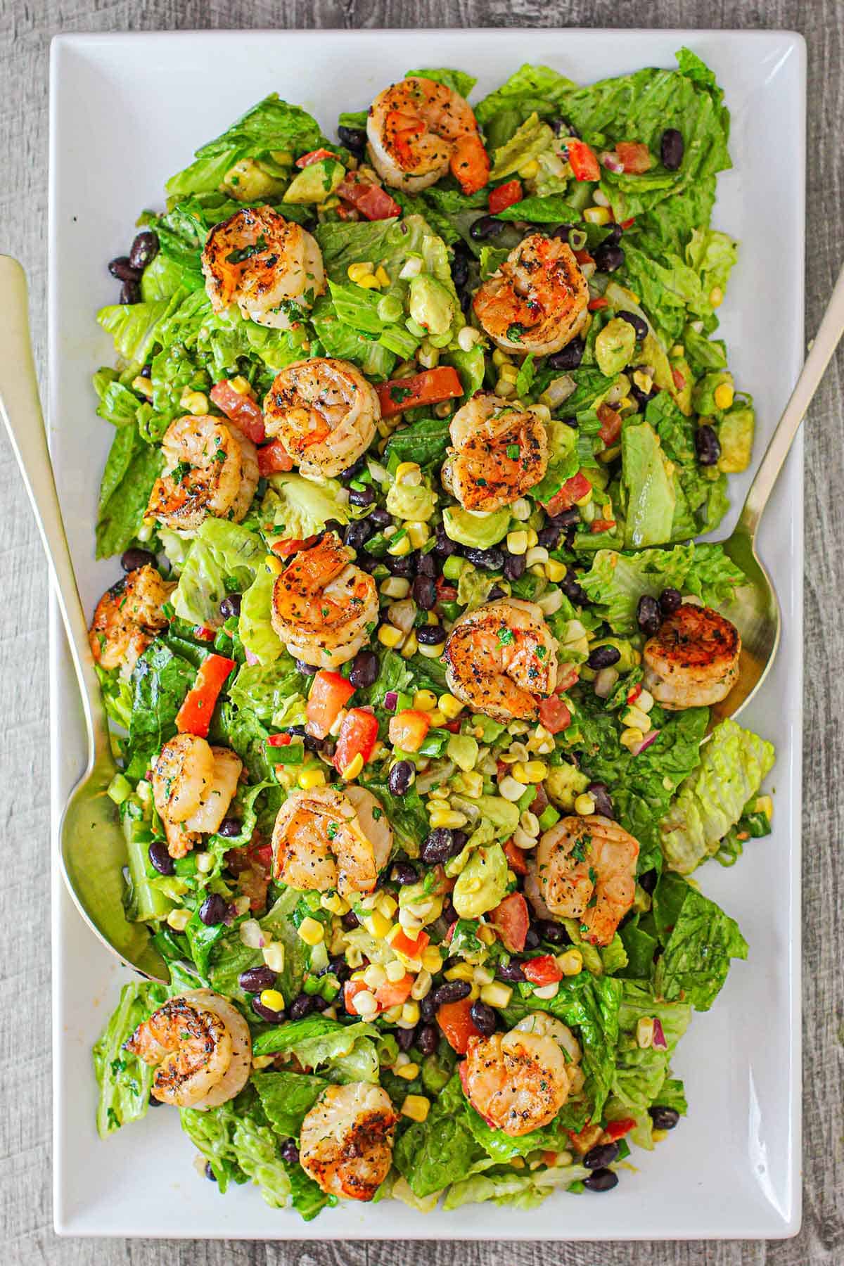 A large rectangular white platter filled with a grilled shrimp salad with cilantro-lime dressing along with grilled corn, black beans, chopped lettuce, all on torn pieces of romaine lettuce.