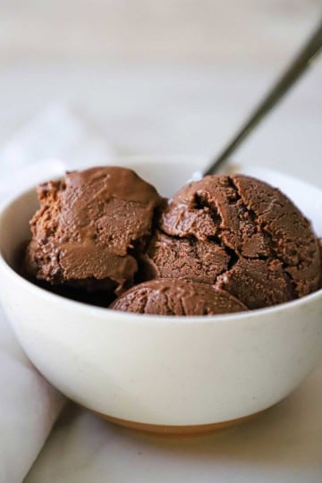 A white ice cream bowl filled with three scoops of homemade chocolate ice cream with a silver spoon inserted off to the side.