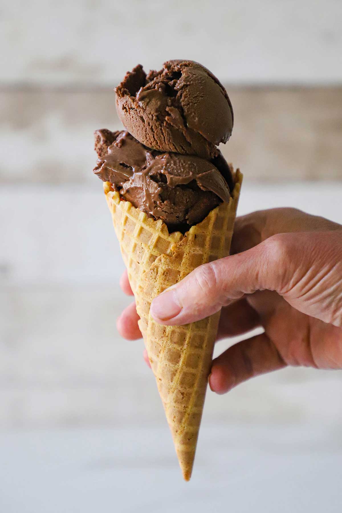 A person holding a waffle cone filled with two scoops of homemade chocolate ice cream.