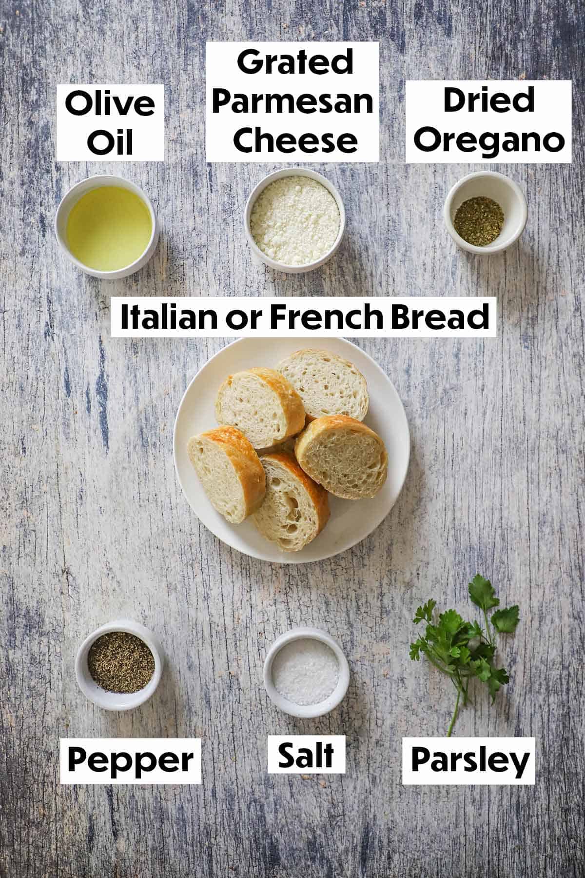 An arrangement of ingredients for homemade croutons on a grey wooden background including sliced bread, olive oil, Parmesan cheese, spices, and fresh parsley.
