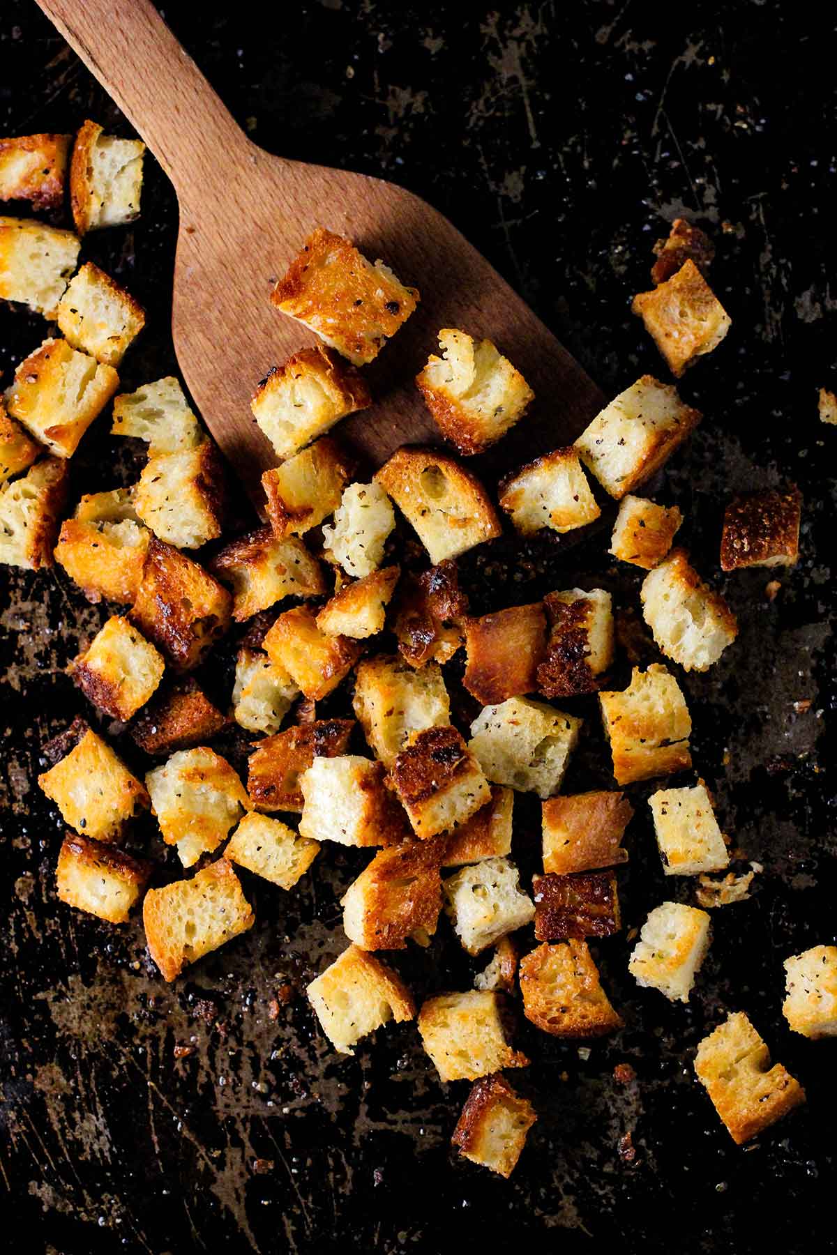 A scratched metal baking sheet filled with homemade croutons with a wooden spatula holding some of the croutons.