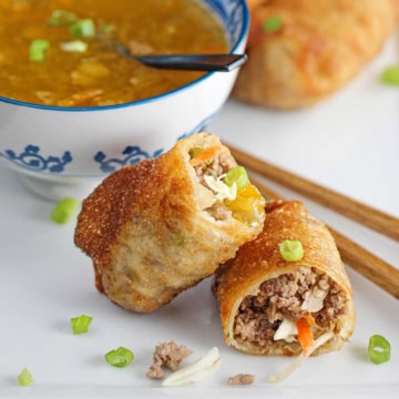 A homemade egg roll that has been split in half revealing the pork and cabbage inside with one of the halves resting on the other half with a bowl of homemade duck sauce in the background.