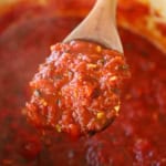 A wooden spoon holding up a large spoonful of homemade marinara sauce over a pot filled with the simmering sauce.