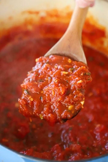 A wooden spoon holding up a large spoonful of homemade marinara sauce over a pot filled with the simmering sauce.
