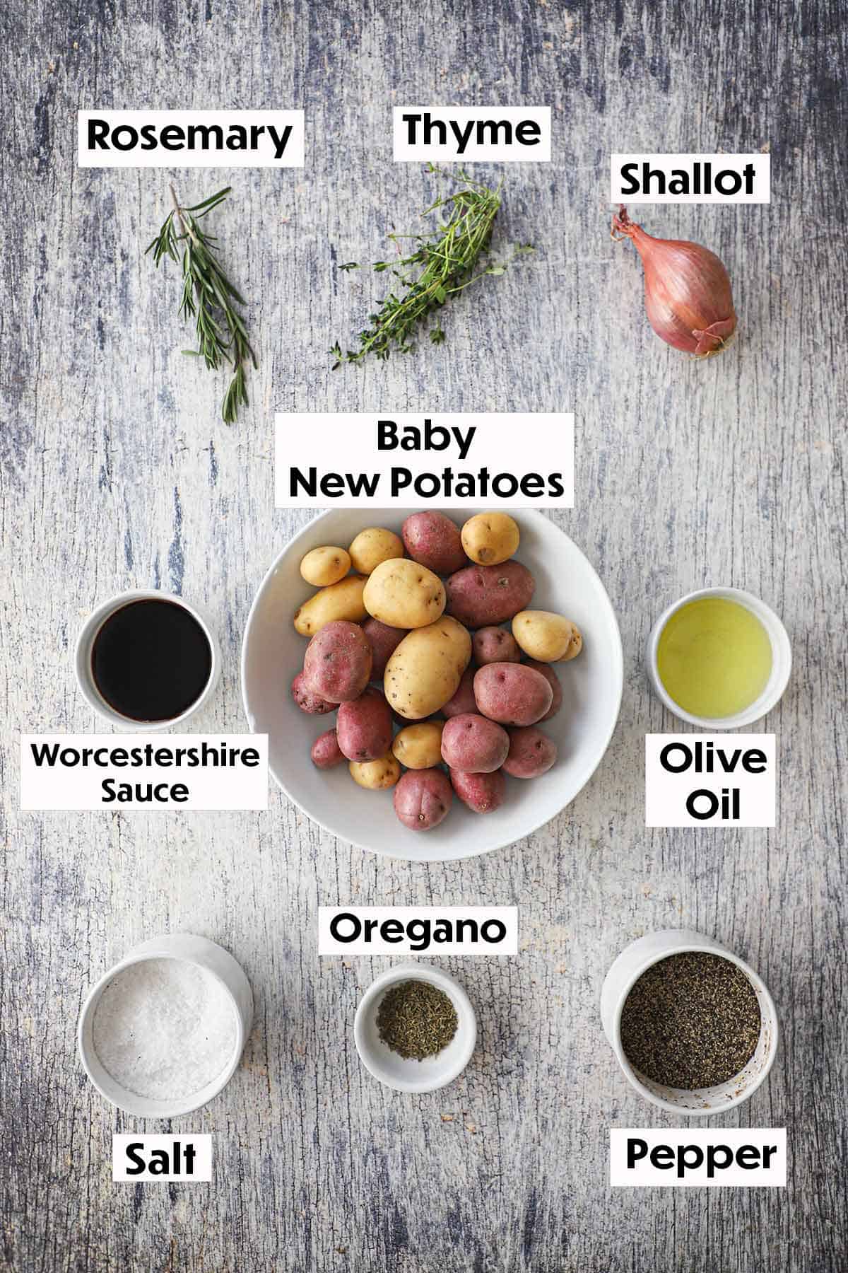 An arrangement of ingredients for roasted potatoes with balsamic and herbs on a wooden grey background including new potatoes, fresh herbs, shallot, Worcestershire sauce, olive oil, dried oregano, salt, and pepper.